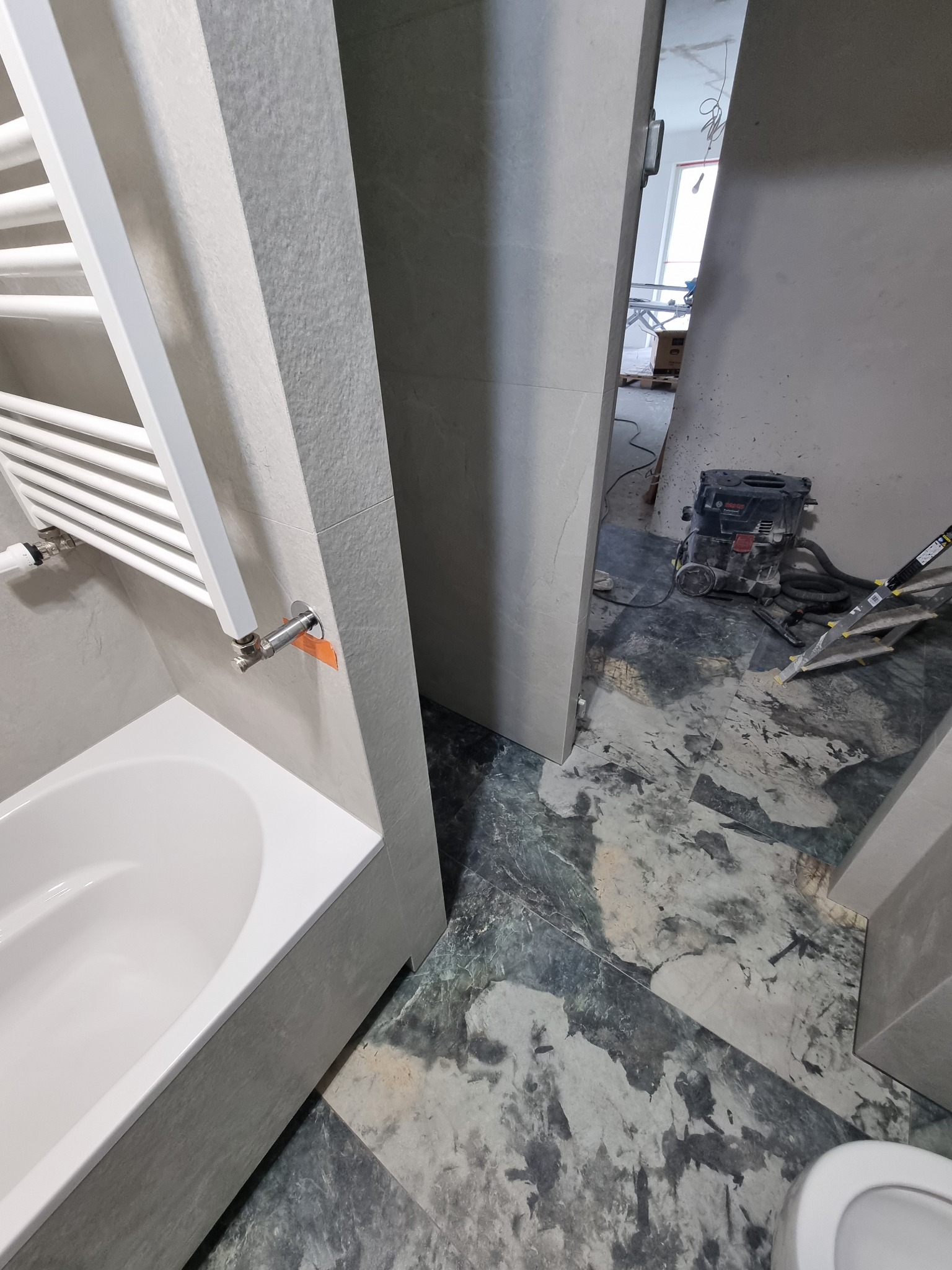 Wnętrze łazienki w trakcie remontu: zamontowana wanna z grzejnikiem drabinkowym, widoczne płytki imitujące marmur na podłodze, odkurzacz przemysłowy i drabina w tle.