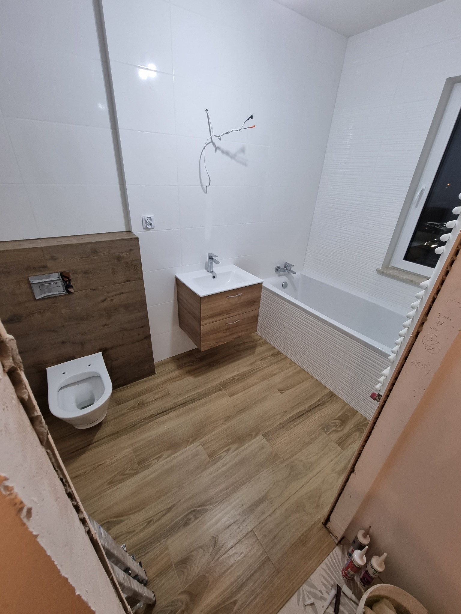 Wnętrze łazienki w trakcie prac wykończeniowych: ułożone płytki imitujące drewno na podłodze, białe płytki na ścianach, wisząca szafka z umywalką, toaleta z zabudową z płyty meblowej oraz widoczna...