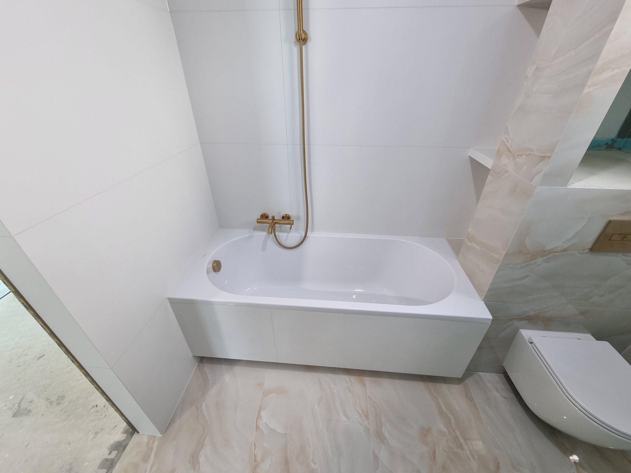 Biała wanna z zabudową, złota armatura, płytki imitujące marmur na ścianie i podłodze, podwieszana toaleta.