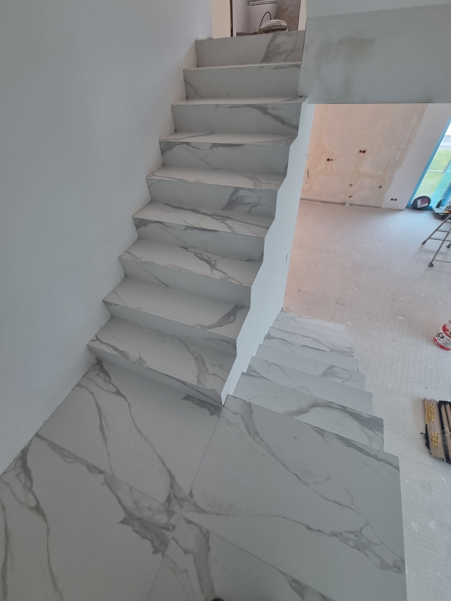 Schody wyłożone białymi płytkami imitującymi marmur, widok z góry, pomieszczenie w trakcie wykańczania.