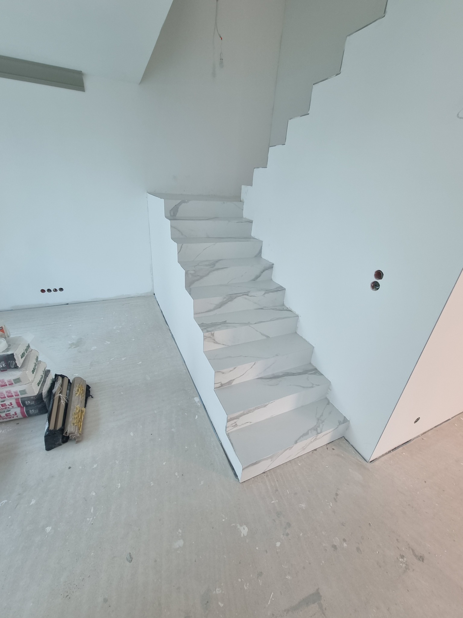 Wykończone schody wewnętrzne obłożone płytkami imitującymi marmur, widoczne worki z klejem i listwy przypodłogowe w niezagospodarowanym wnętrzu.