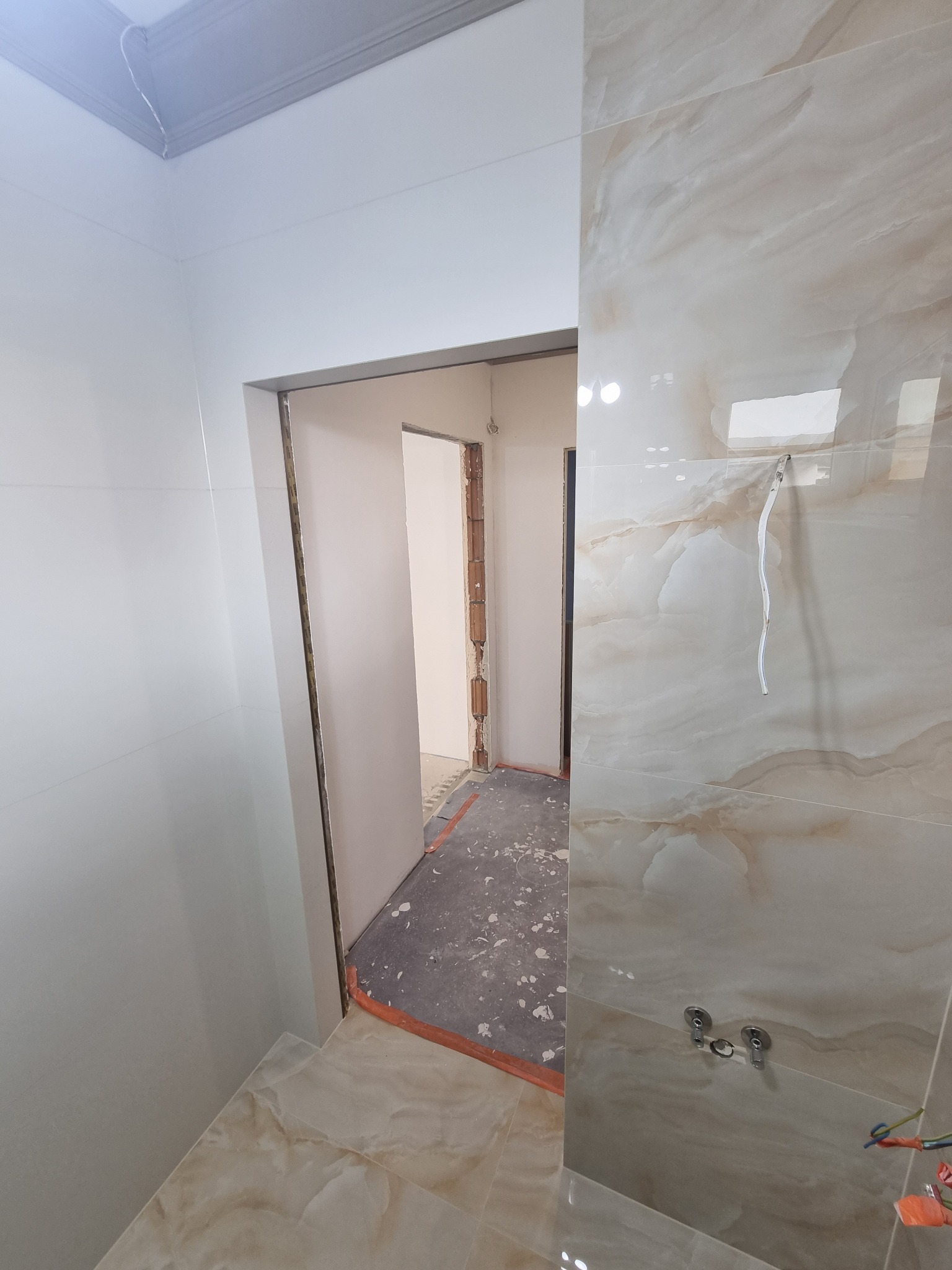 Wnętrze łazienki w trakcie wykańczania: beżowe płytki imitujące marmur na ścianach i podłodze, widoczne przyłącza wodne i wystające kable, surowe przejście drzwiowe z widocznym fragmentem muru...