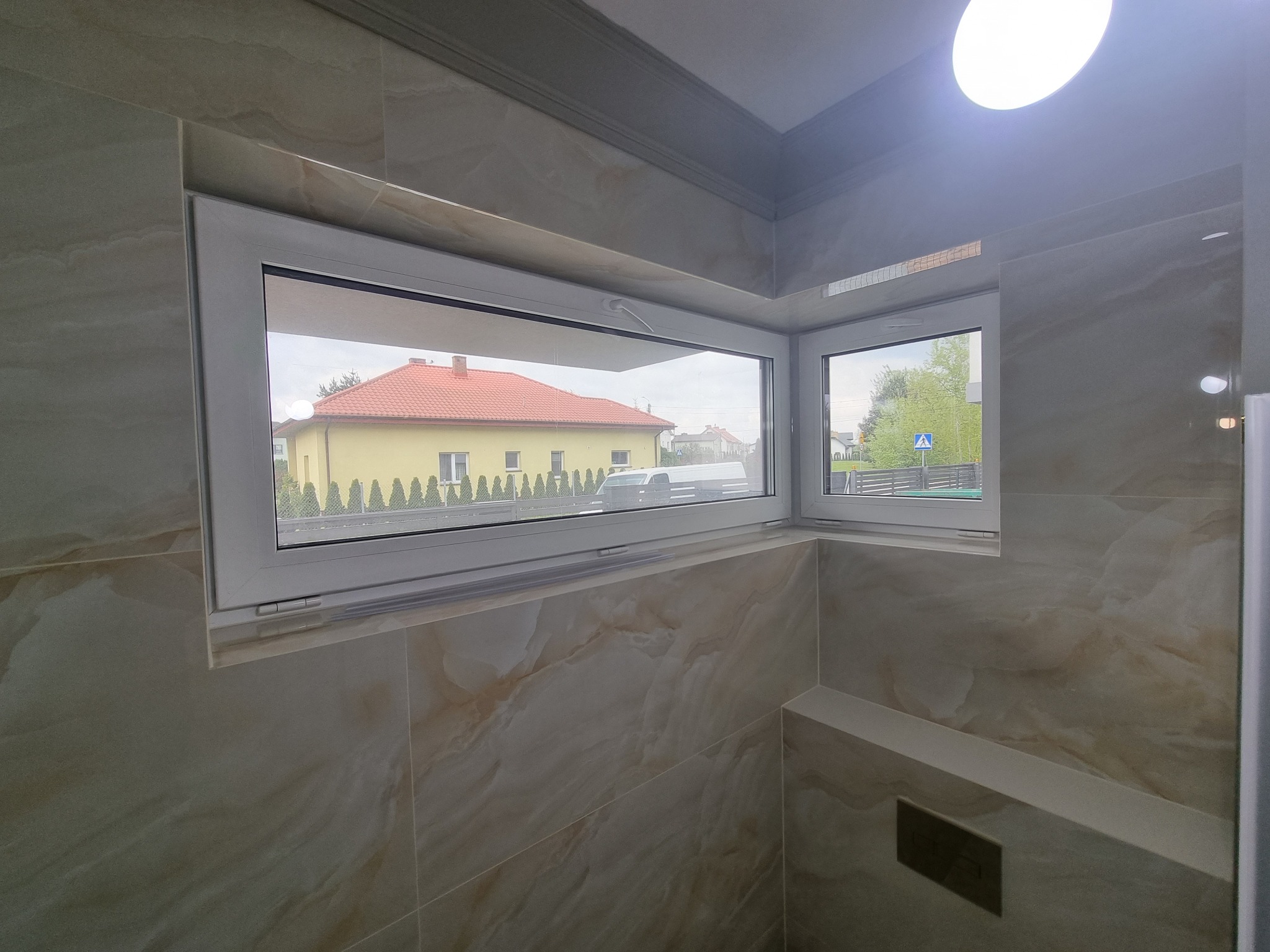 Beżowe płytki imitujące marmur na ścianach w łazience w Warszawie, widok przez okno na dom z czerwonym dachem.