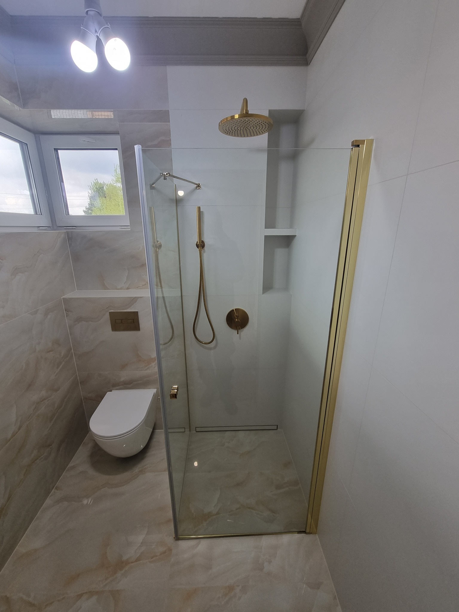 Nowoczesna łazienka z kabiną prysznicową typu walk-in z złotymi profilami, deszczownicą i baterią w kolorze złota, marmurowe płytki na podłodze i ścianach, wisząca toaleta.
