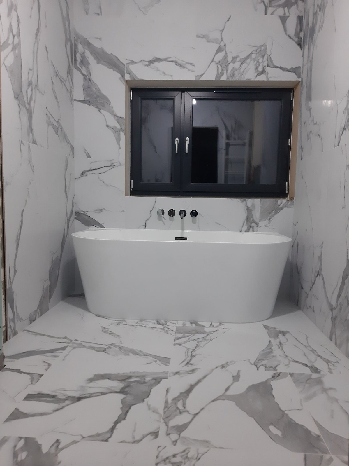 Nowoczesna łazienka z wolnostojącą białą wanną, wykończona płytkami imitującymi marmur, z czarnym oknem i minimalistyczną armaturą.