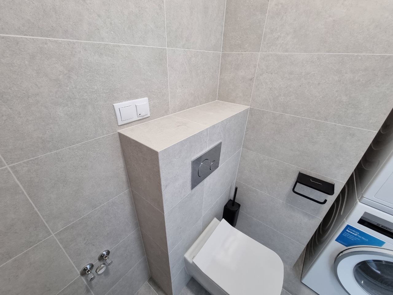 Szara łazienka z podtynkową toaletą, zabudową z płytek, i pralką Bosch SpeedPerfect w rogu.