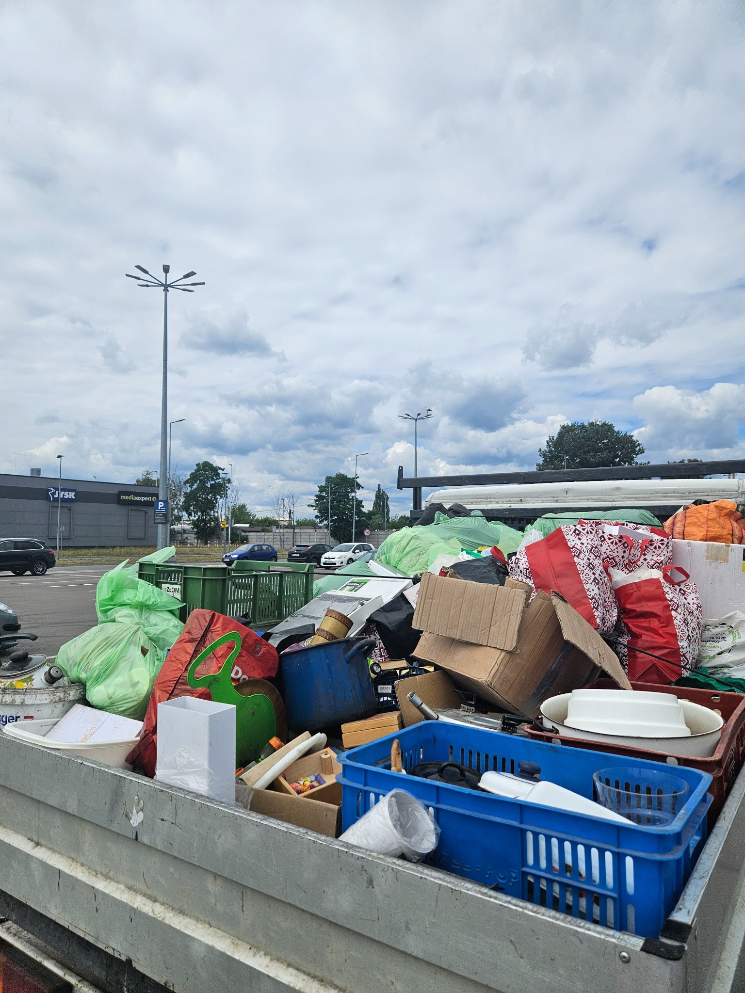 Pełna skrzynia załadowana różnorodnymi przedmiotami, w tym torby, pudełka kartonowe i plastikowe pojemniki, na tle parkingu z budynkiem sklepowym w pochmurny dzień.