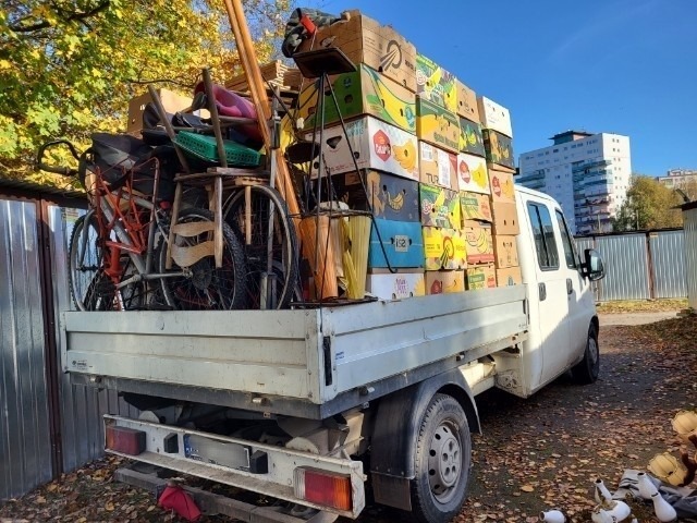 Tył białej ciężarówki załadowanej po brzegi kartonami z bananami, rowerami, krzesłami i innymi przedmiotami podczas transportu, na tle bloku mieszkalnego i garaży.