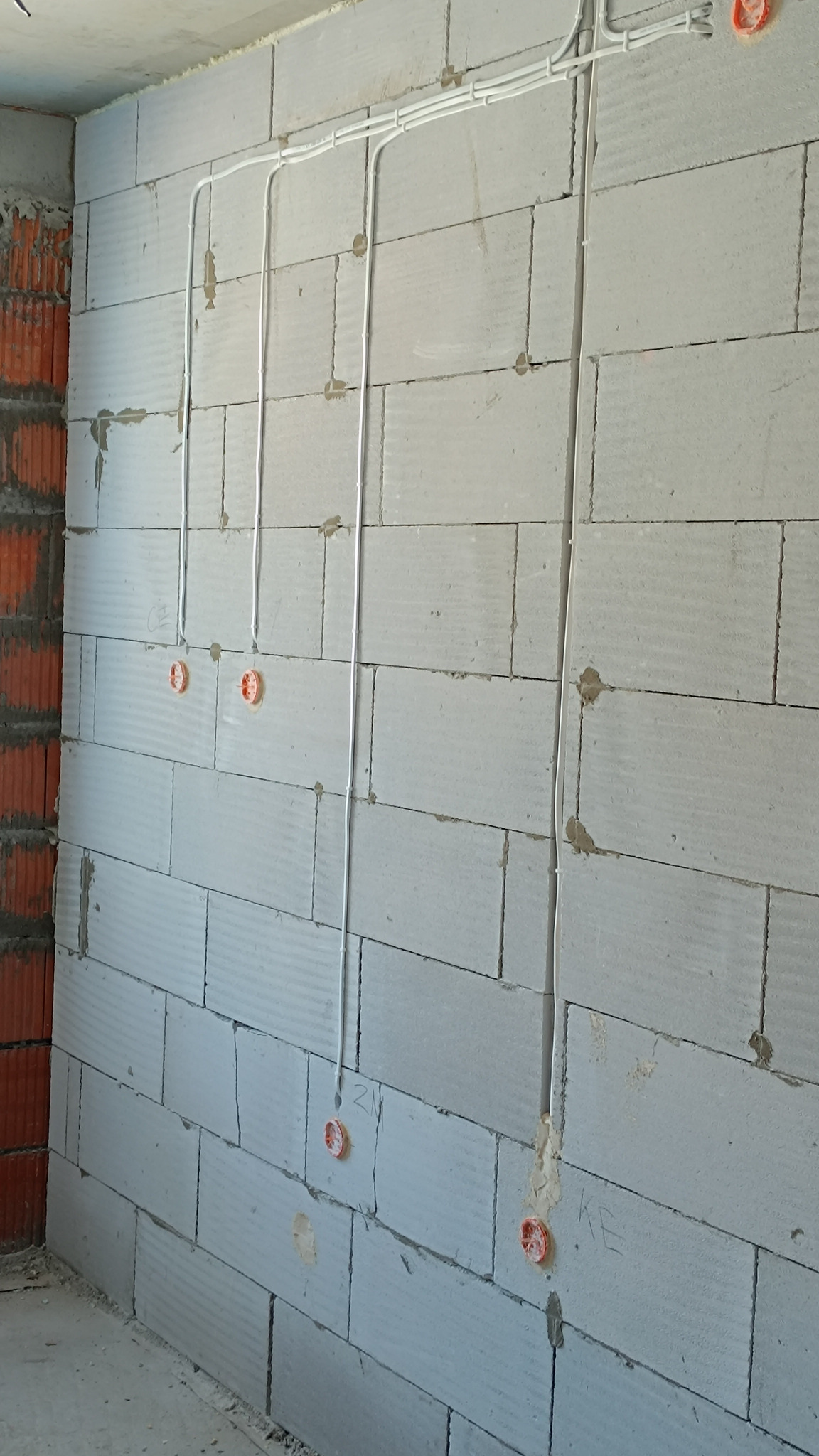 Białe przewody elektryczne ułożone w bruzdach w ścianie z białych bloczków, widoczne pomarańczowe puszki instalacyjne, ściana w stanie surowym.