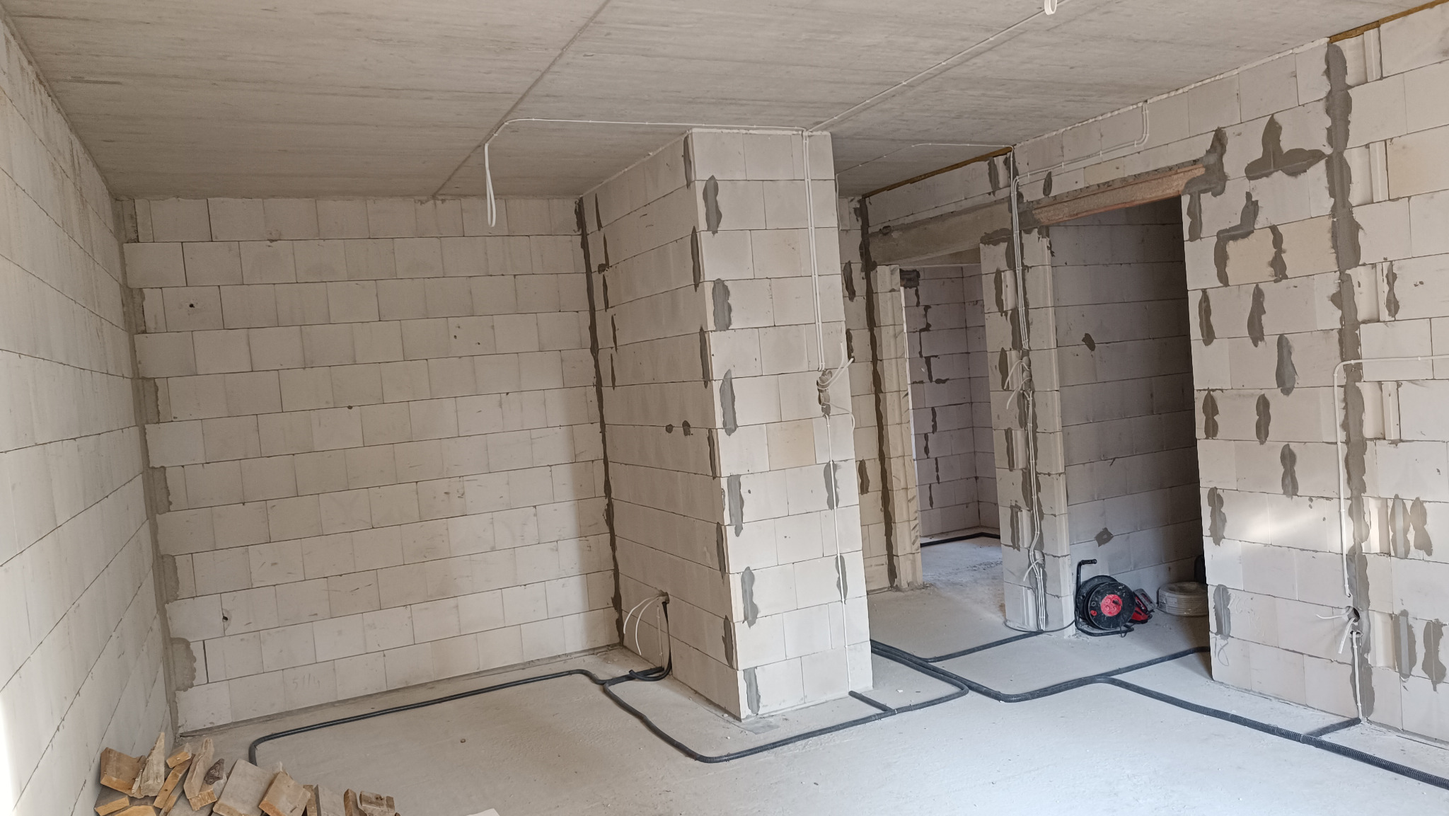 Surowy stan wnętrza z widocznymi przewodami elektrycznymi w peszlach przy podłodze i na ścianach, przed tynkowaniem.