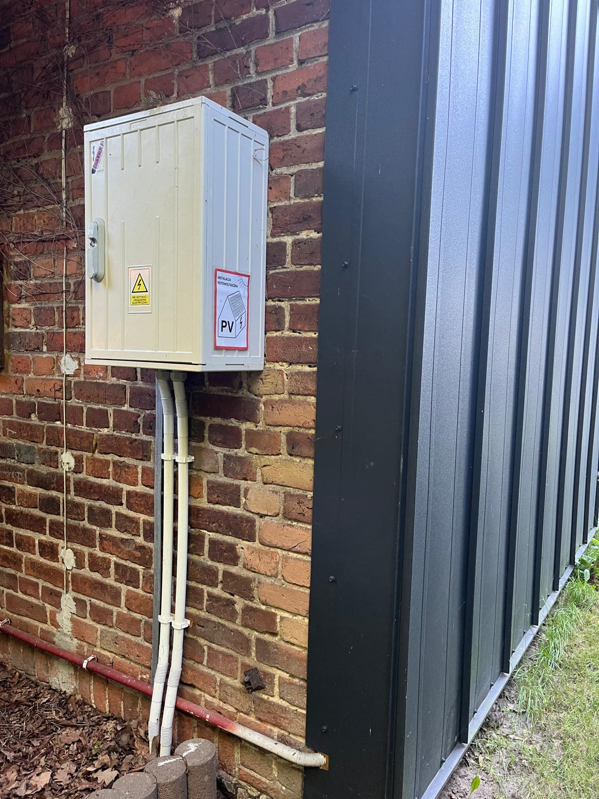 Zewnętrzna skrzynka elektryczna z naklejką instalacji fotowoltaicznej, zamontowana na ceglanej ścianie budynku, z widocznymi rurami osłonowymi przewodów.