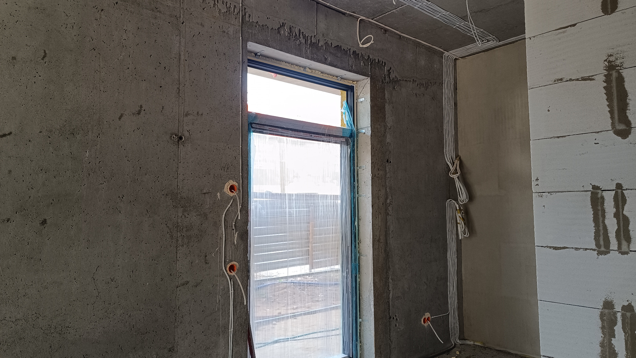 Surowy betonowy pokój w trakcie budowy z widocznymi kablami elektrycznymi wychodzącymi ze ścian, okno zabezpieczone folią ochronną, a na suficie ułożone przewody w korytkach.