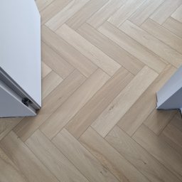 MARTIN Marcin Kłosowski - Jasne panele ułożone w jodełkę, widok z góry z fragmentem białych ościeżnic drzwiowych po bokach i butami w dolnej części kadru.