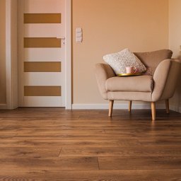 MARTIN Marcin Kłosowski - Beżowy fotel z poduszką i filiżanką na złotej tacy, na drewnianej podłodze, obok białych drzwi z poziomymi, złotymi paskami w pokoju o ciepłych, beżowych ścianach.