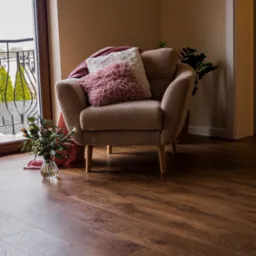 Ułożona podłoga z paneli imitujących drewno z widocznym fotelem, poduszkami i bukietem kwiatów w wazonie przy oknie balkonowym.