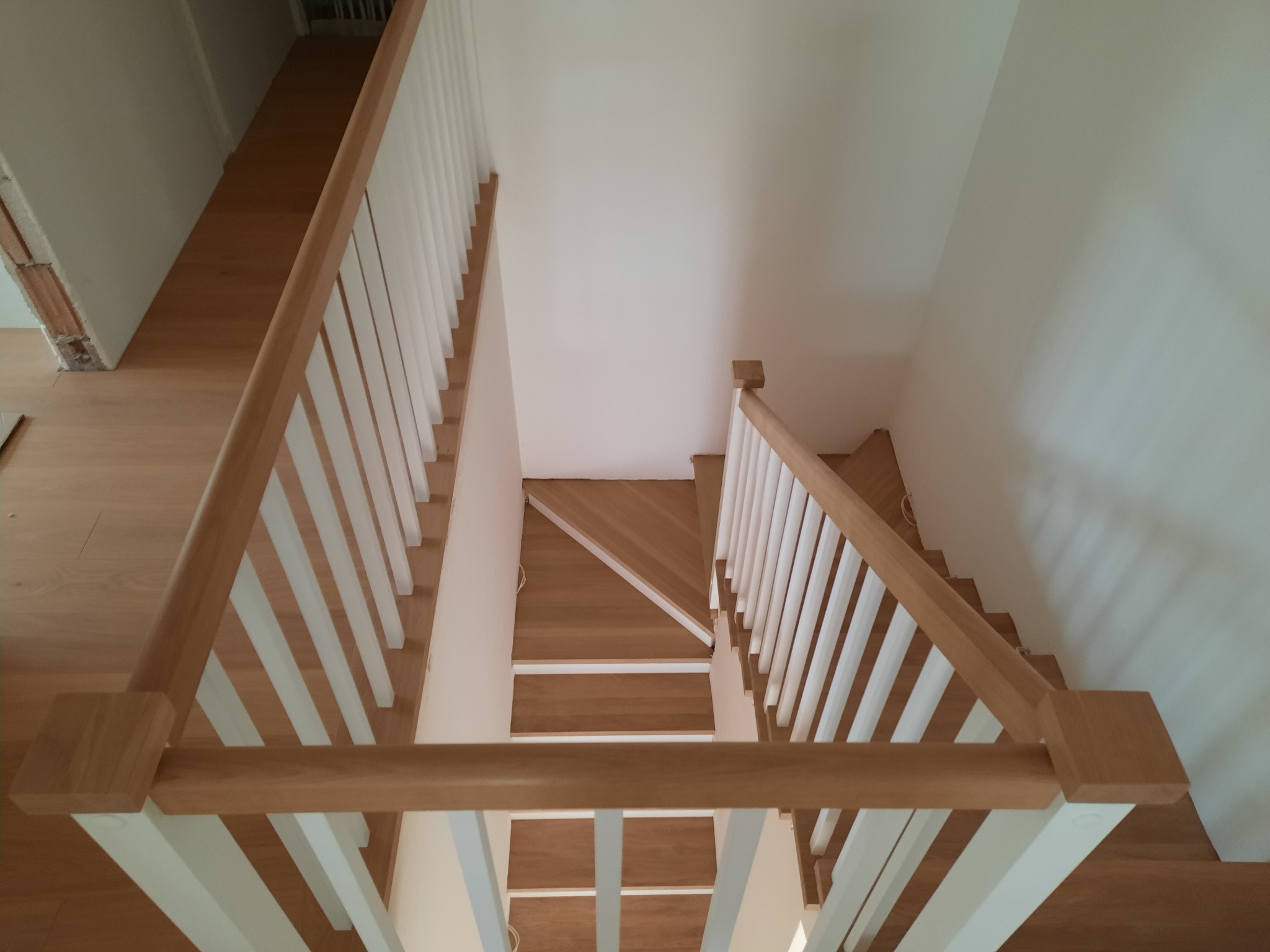 Widok z góry na drewniane schody z białymi balustradami, prowadzące w dół, z widocznymi panelami na podłodze i białymi ścianami.