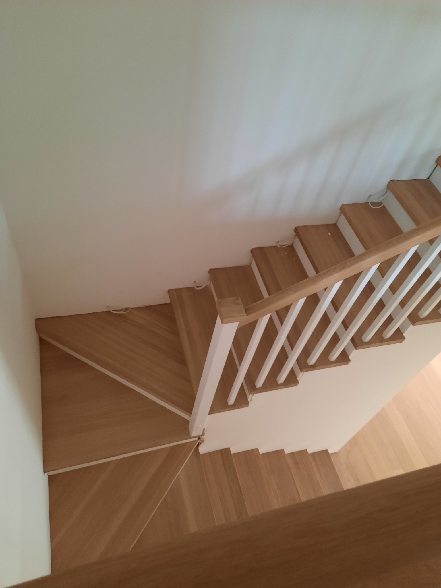 Widok z góry na drewniane schody z białymi balustradami, z widocznymi niedoskonałościami wykończenia przy ścianie i wystającymi kablami.