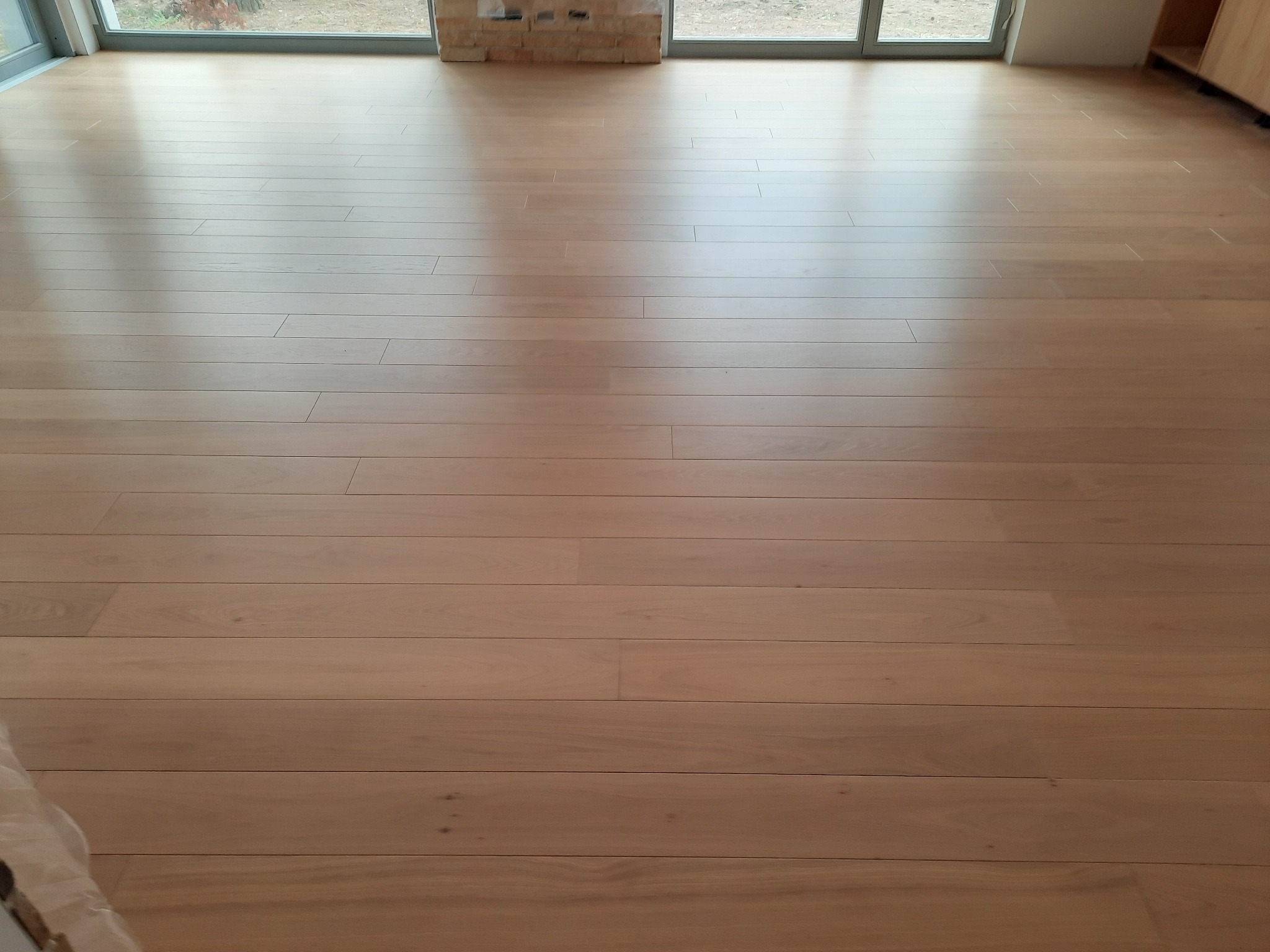 Jasna, nowa podłoga z drewnianych paneli w przestronnym pokoju z dużymi oknami, z widocznym naturalnym światłem i fragmentem ściany z cegły w tle.