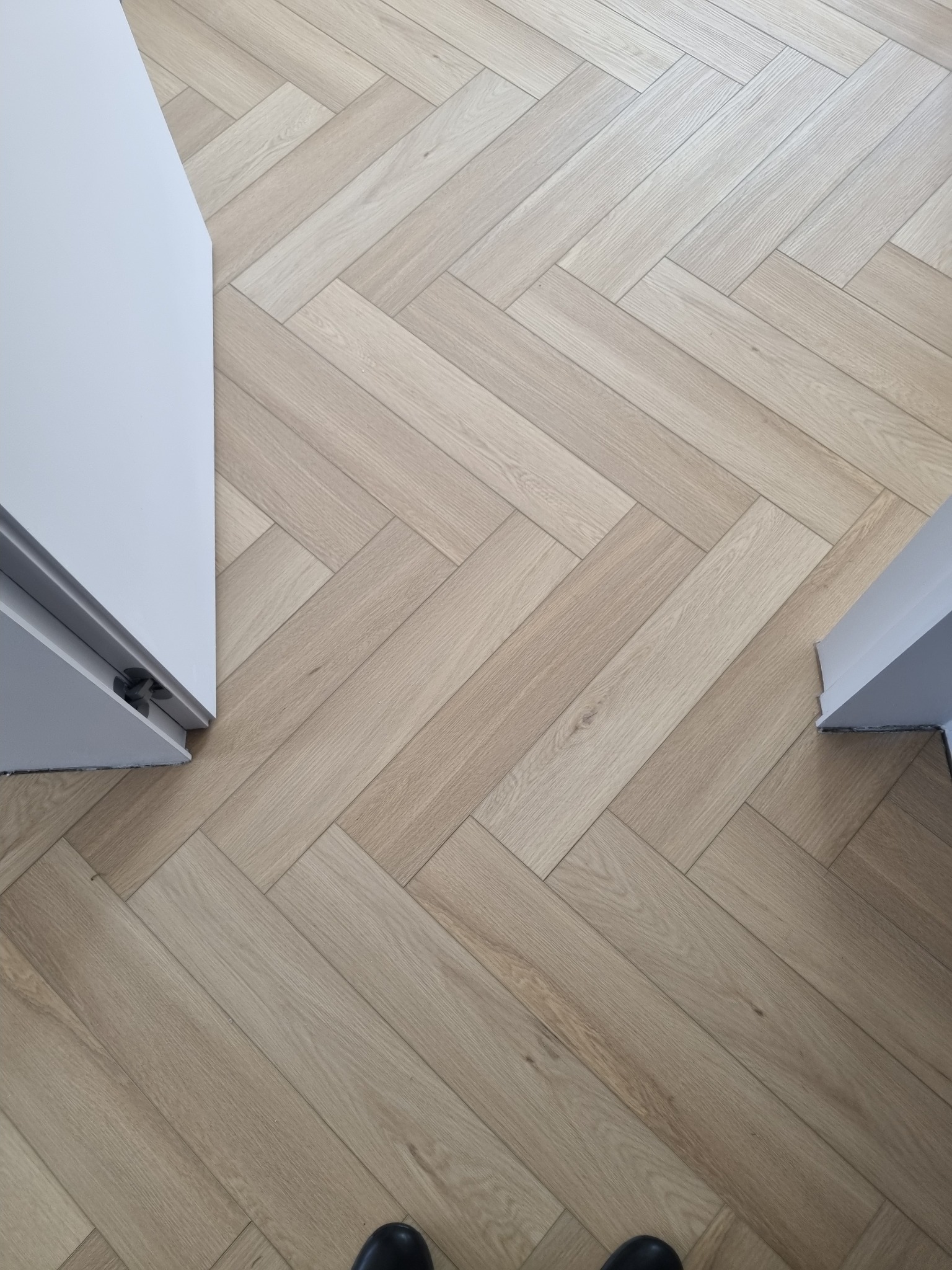 Jasne panele ułożone w jodełkę, widok z góry z fragmentem białych ościeżnic drzwiowych po bokach i butami w dolnej części kadru.