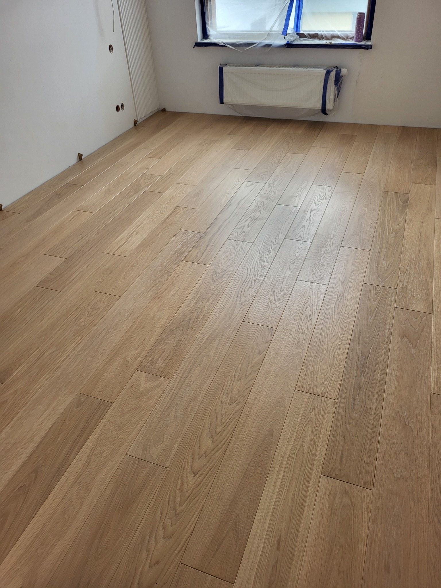 Ułożona jasna podłoga z paneli w nowym mieszkaniu w Warszawie, z zabezpieczonym folią oknem i grzejnikiem.