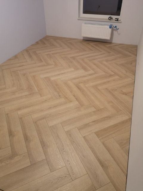 Jasne panele ułożone w jodełkę francuską, widok na fragment pokoju z grzejnikiem pod oknem.