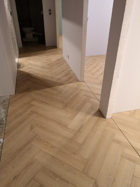 Jasny parkiet ułożony we wzór jodełki w przejściu między pokojami, widoczne niedokończone wykończenia ścian i surowy stan futryn drzwiowych.