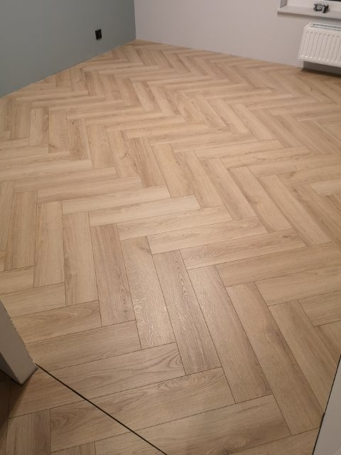 Ułożona jasna podłoga z paneli w jodełkę francuską, widok z niskiej perspektywy, fragment ściany w kolorze gołębim i biały grzejnik pod oknem w tle.