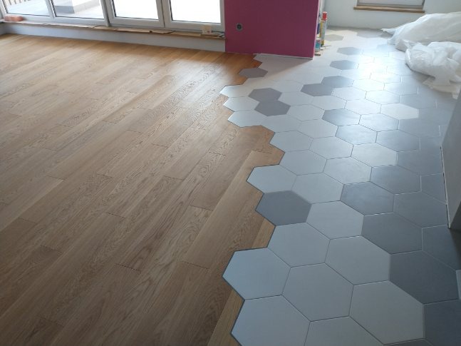 Fragment podłogi z widocznym połączeniem parkietu i heksagonalnych płytek w odcieniach szarości, widoczne okno i różowy fragment ściany