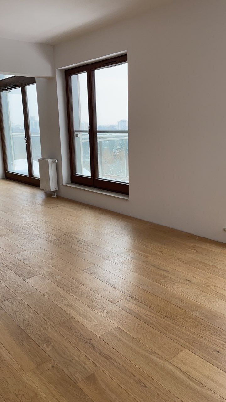 Jasna, drewniana podłoga ułożona w nowym mieszkaniu z widokiem na miasto przez duże okna z brązowymi ramami i białym grzejnikiem pod jednym z nich.
