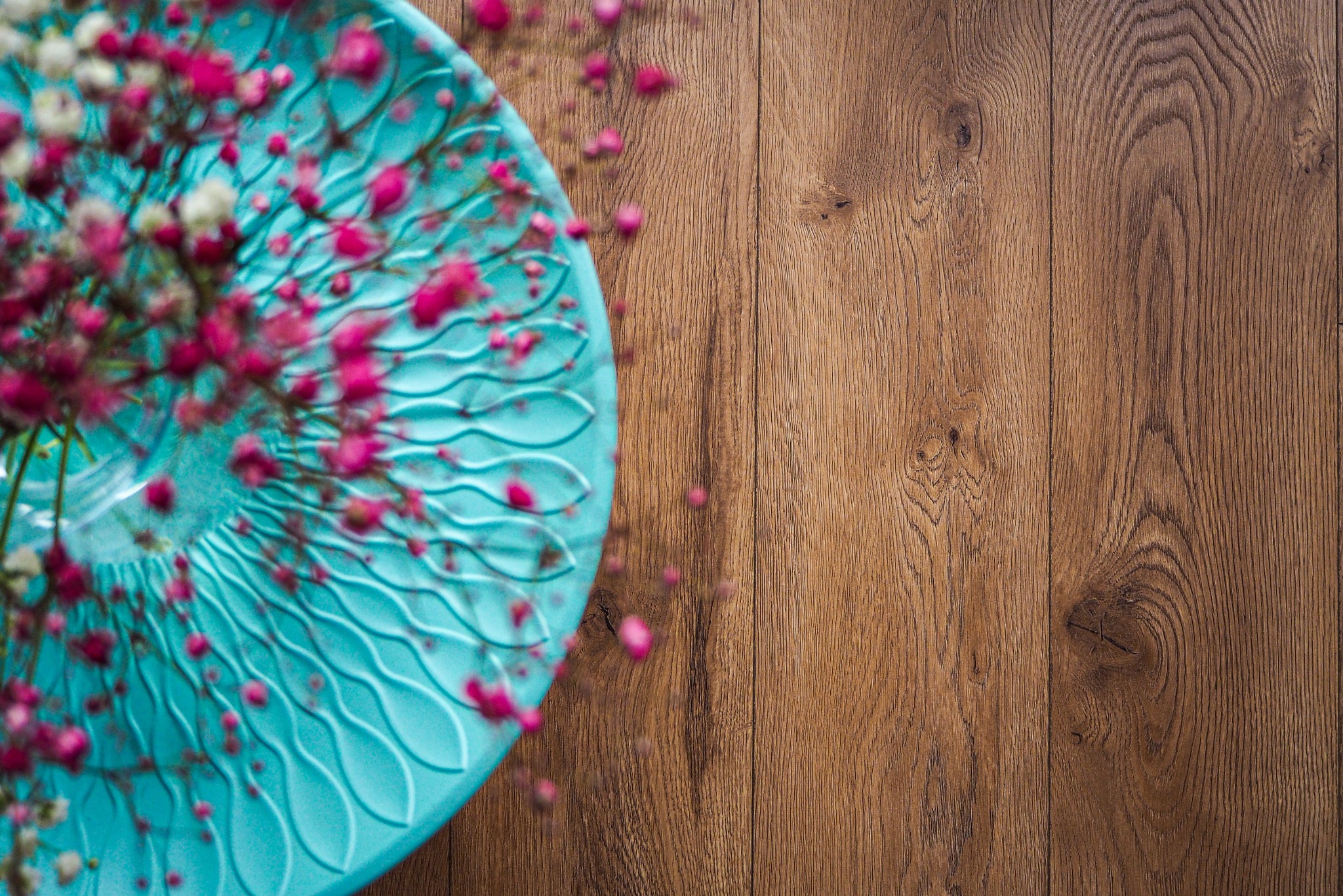 Fragment drewnianej podłogi z dekoracyjnym turkusowym talerzem i drobnymi różowymi kwiatami, ujęcie z góry.