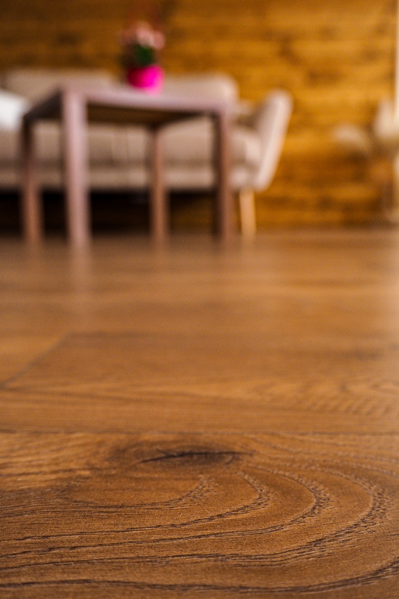 Zbliżenie na nową, brązową podłogę z paneli z widocznym wzorem drewna, w tle rozmyty stół z dekoracją kwiatową i drewniana ściana.