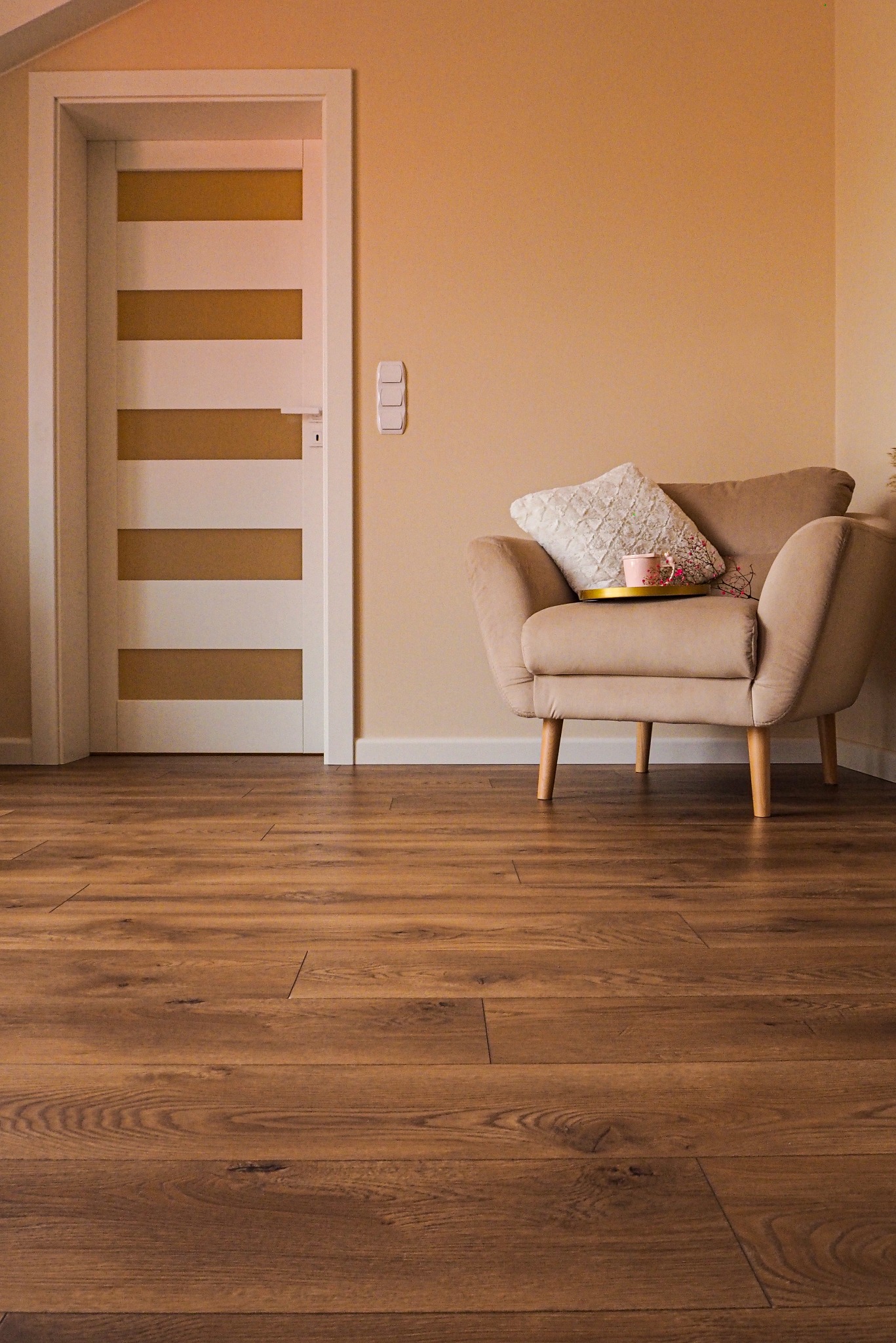 Beżowy fotel z poduszką i filiżanką na złotej tacy, na drewnianej podłodze, obok białych drzwi z poziomymi, złotymi paskami w pokoju o ciepłych, beżowych ścianach.