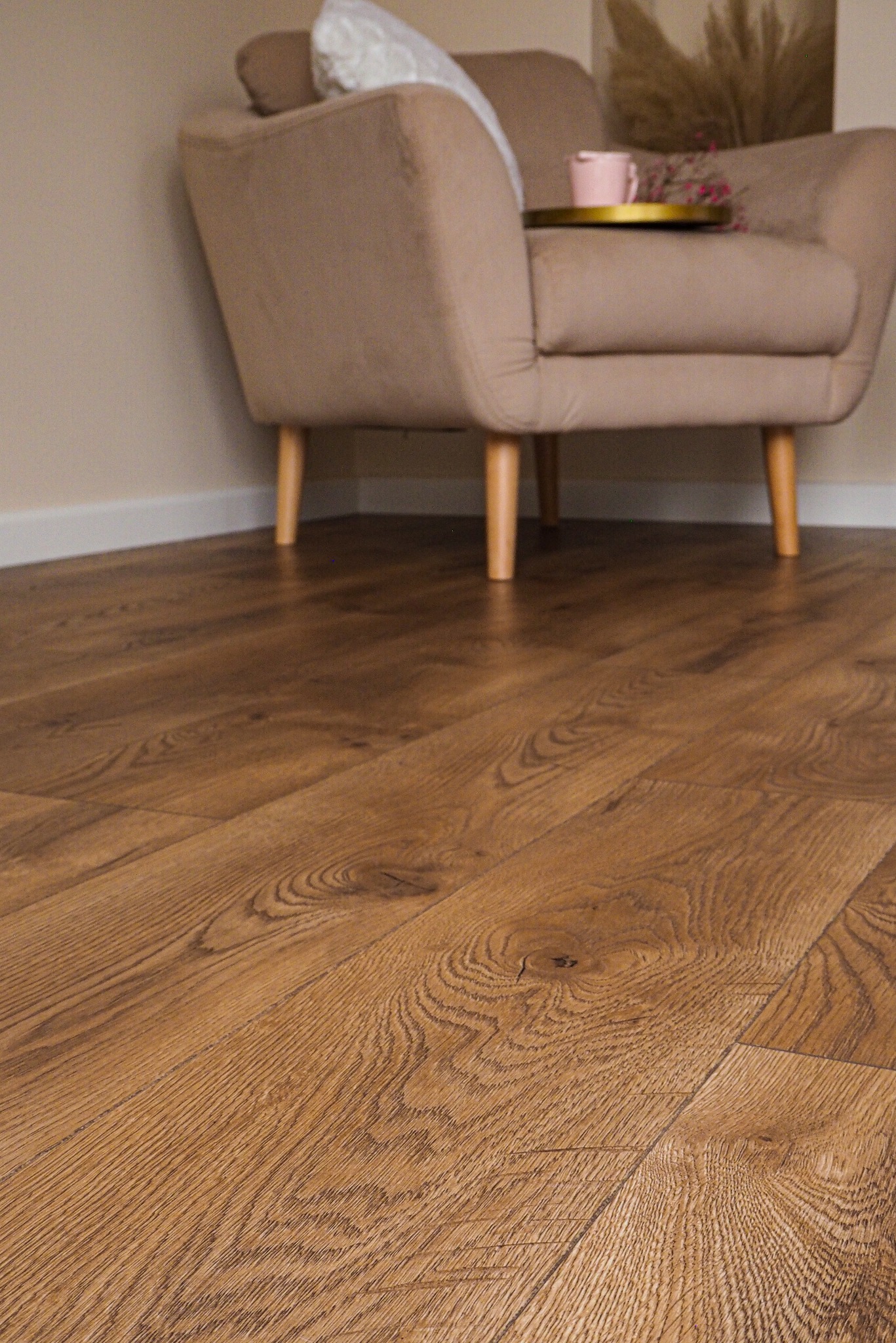 Ukośne ujęcie brązowych paneli podłogowych z widoczną strukturą drewna, w tle fragment beżowego fotela na drewnianych nóżkach z dekoracyjną tacą.
