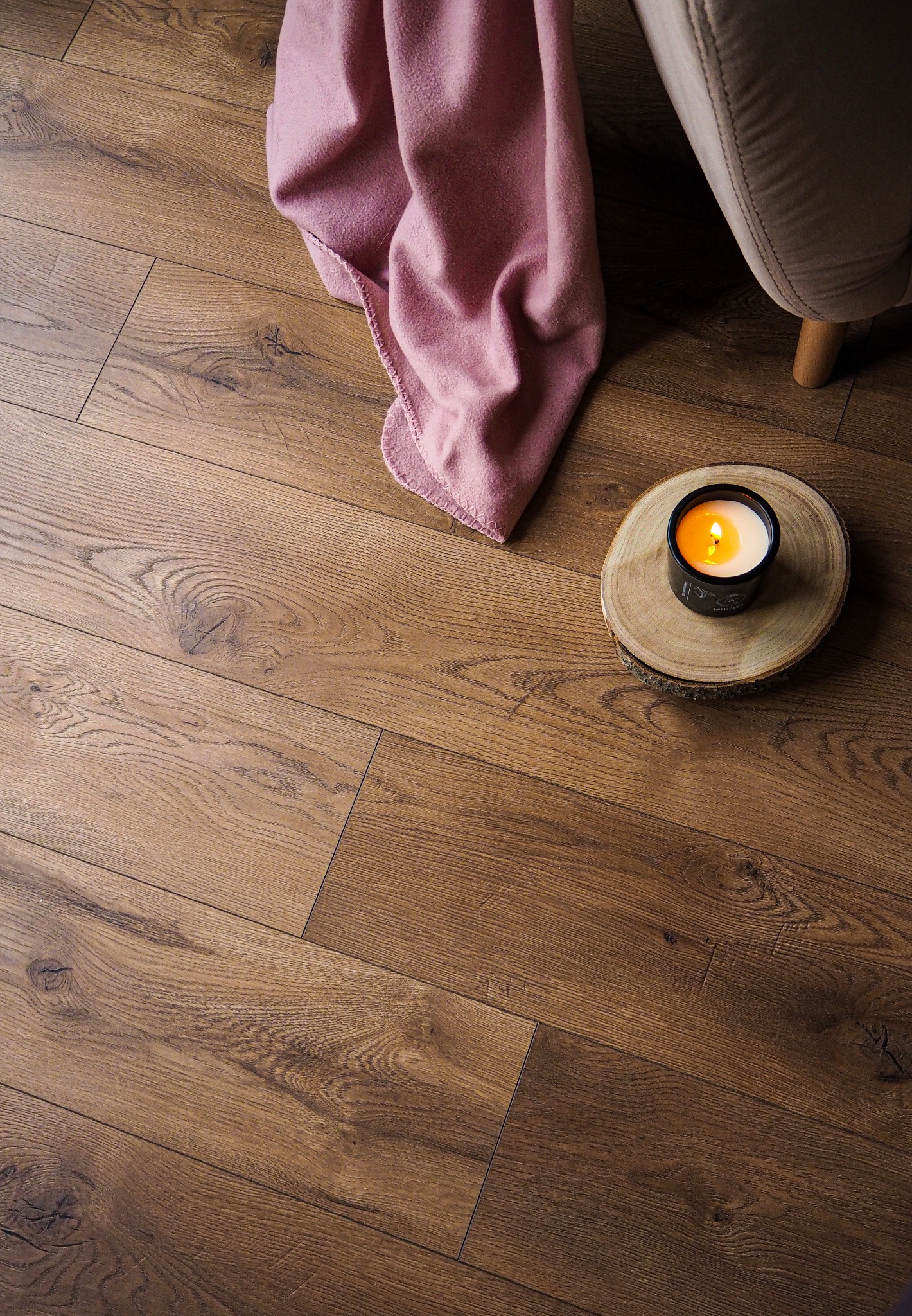 Ułożone panele podłogowe w odcieniach brązu, z widoczną strukturą drewna, uzupełnione o dekoracyjny akcent w postaci zapalonej świecy na drewnianym plastrze i różowego koca, częściowo widoczny...