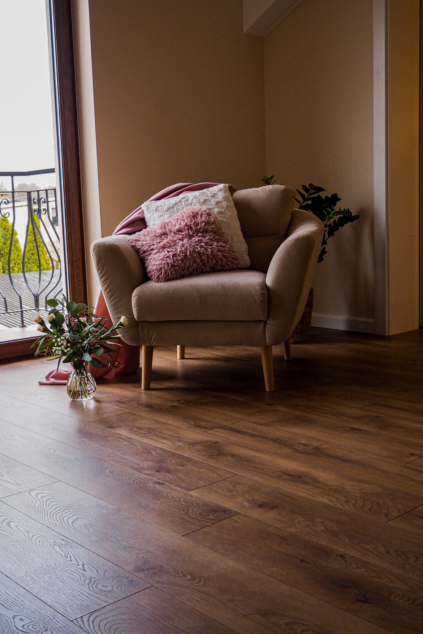 Ułożona podłoga z paneli imitujących drewno z widocznym fotelem, poduszkami i bukietem kwiatów w wazonie przy oknie balkonowym.