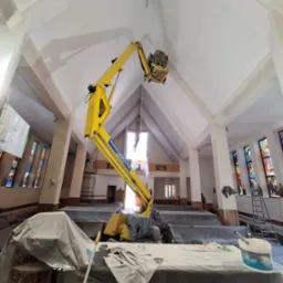 Wnętrze kościoła w trakcie prac remontowych, widoczny podnośnik koszowy z pracownikiem przy suficie, zabezpieczone ławki i sprzęt malarski.