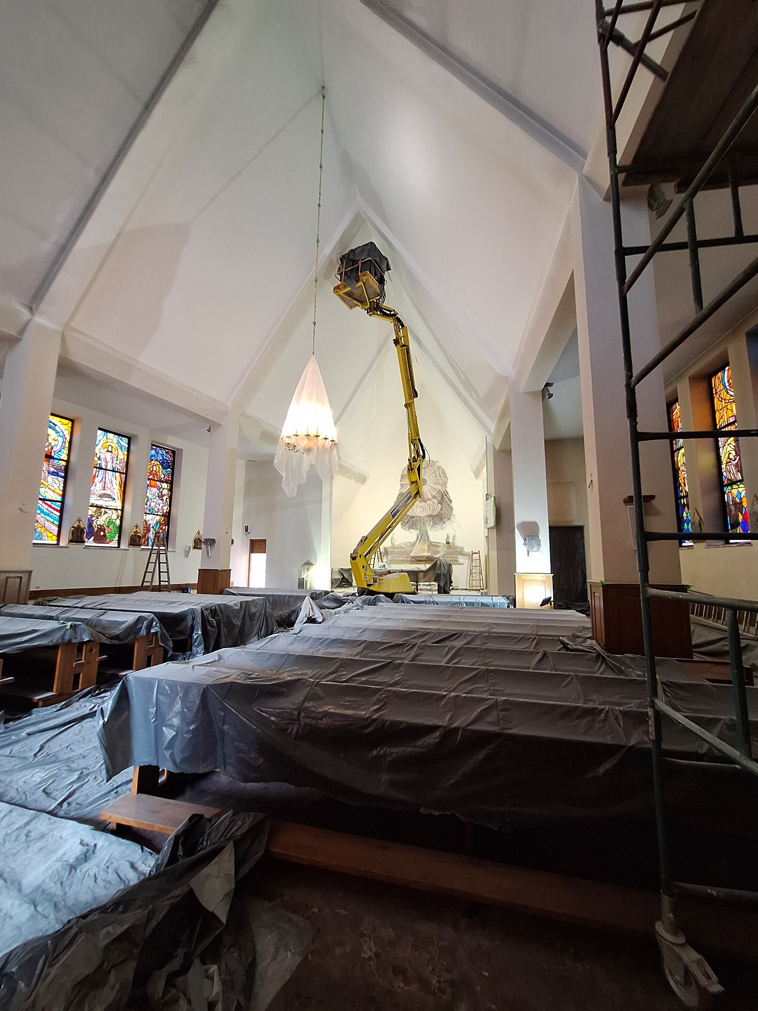 Wnętrze kościoła z wysokim sklepieniem w trakcie prac, zabezpieczone ławki pod folią, podnośnik koszowy w centralnej części, widoczne rusztowania i witraże.