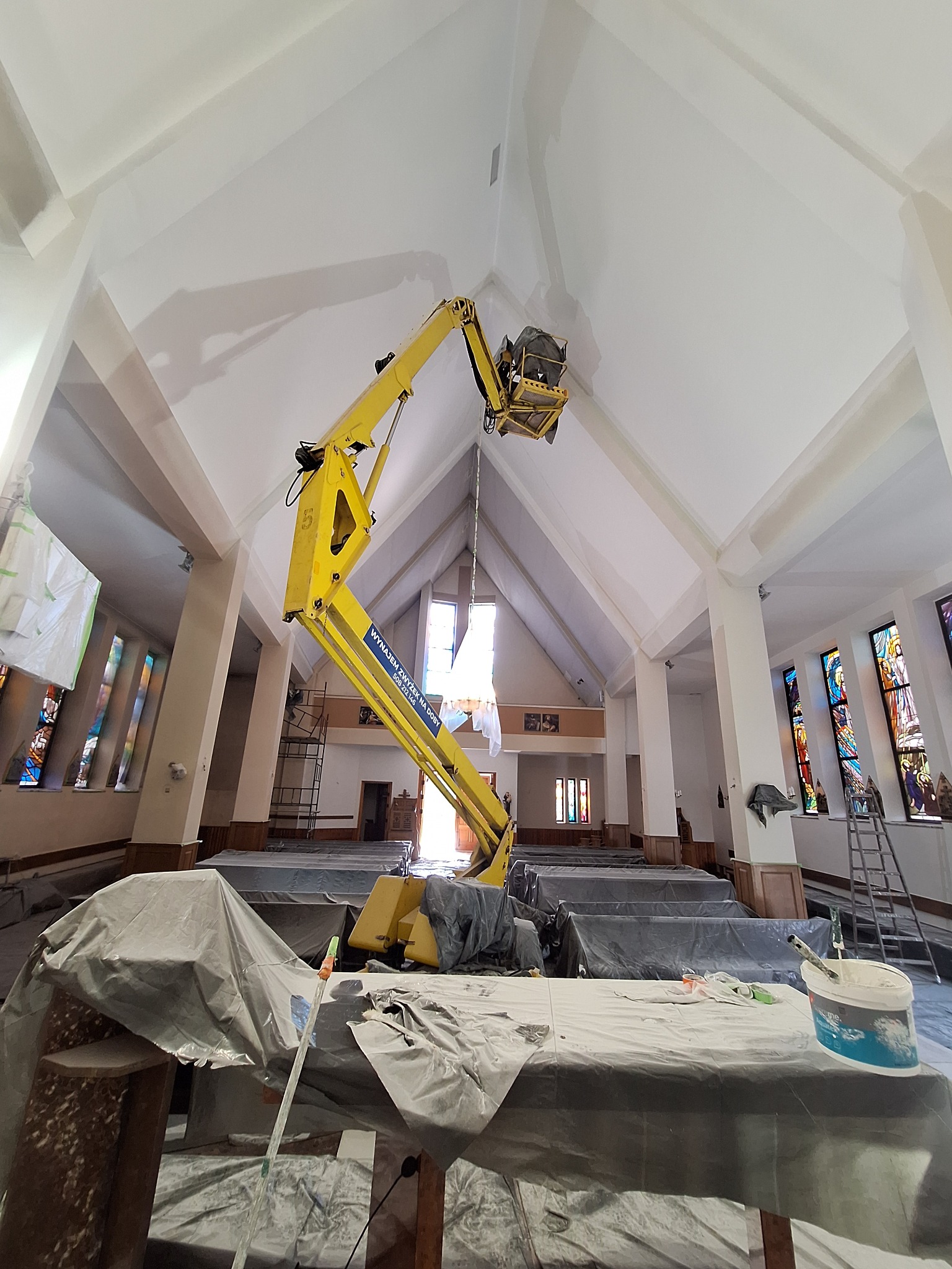 Wnętrze kościoła w trakcie prac remontowych, widoczny podnośnik koszowy z pracownikiem przy suficie, zabezpieczone ławki i sprzęt malarski.