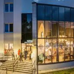 Nowoczesny budynek biurowy z przeszkloną fasadą i oświetlonym wnętrzem. Kilka osób wchodzi po schodach do wejścia. Wieczorny kadr z zielenią w tle.