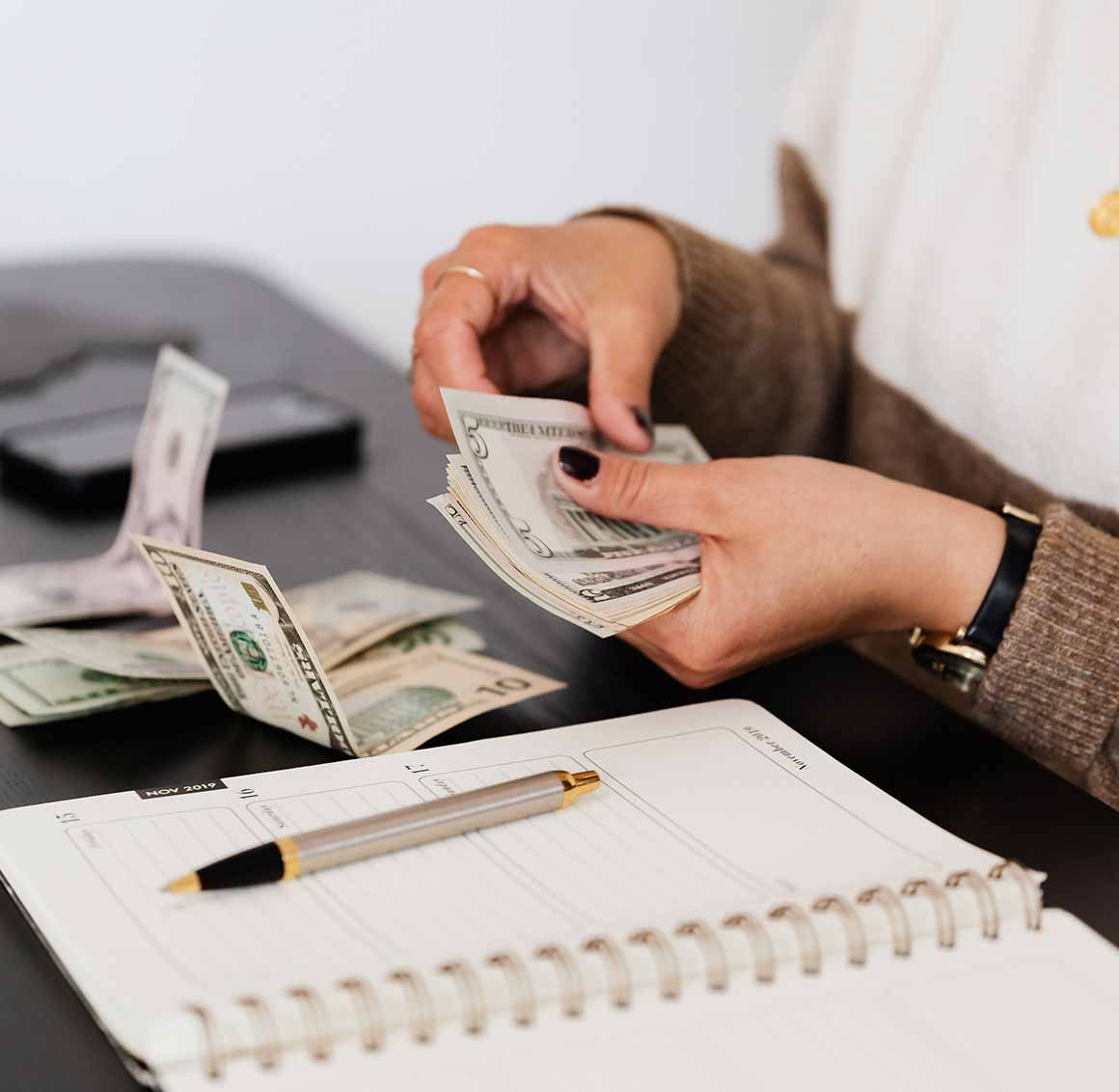Osoba liczy banknoty USD nad kalendarzem i długopisem na biurku. Widoczny telefon. Kadr biurowy, praca z finansami, planowanie wydatków, zarządzanie budżetem.