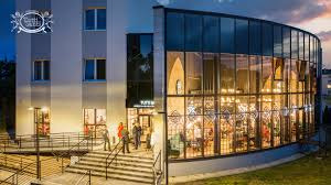 Nowoczesny budynek biurowy z przeszkloną fasadą i oświetlonym wnętrzem. Kilka osób wchodzi po schodach do wejścia. Wieczorny kadr z zielenią w tle.