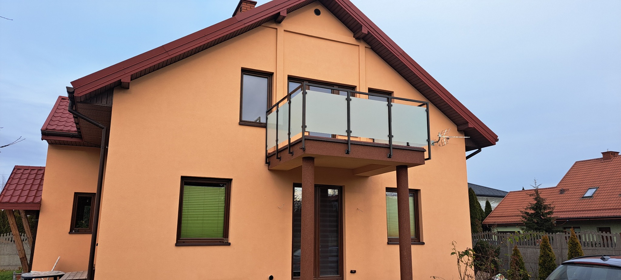 Dom z balkonem zabezpieczonym szklaną konstrukcją na metalowym stelażu, wspartym na dwóch kolumnach. Widoczne brązowe ramy okienne i pomarańczowa elewacja budynku.
