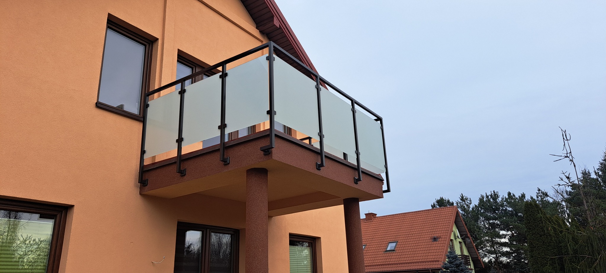 Zewnętrzny balkon z czarną ramą i mlecznym szkłem, widoczny z dołu, z fragmentem elewacji budynku w kolorze brzoskwiniowym i dachem sąsiedniego domu w tle.