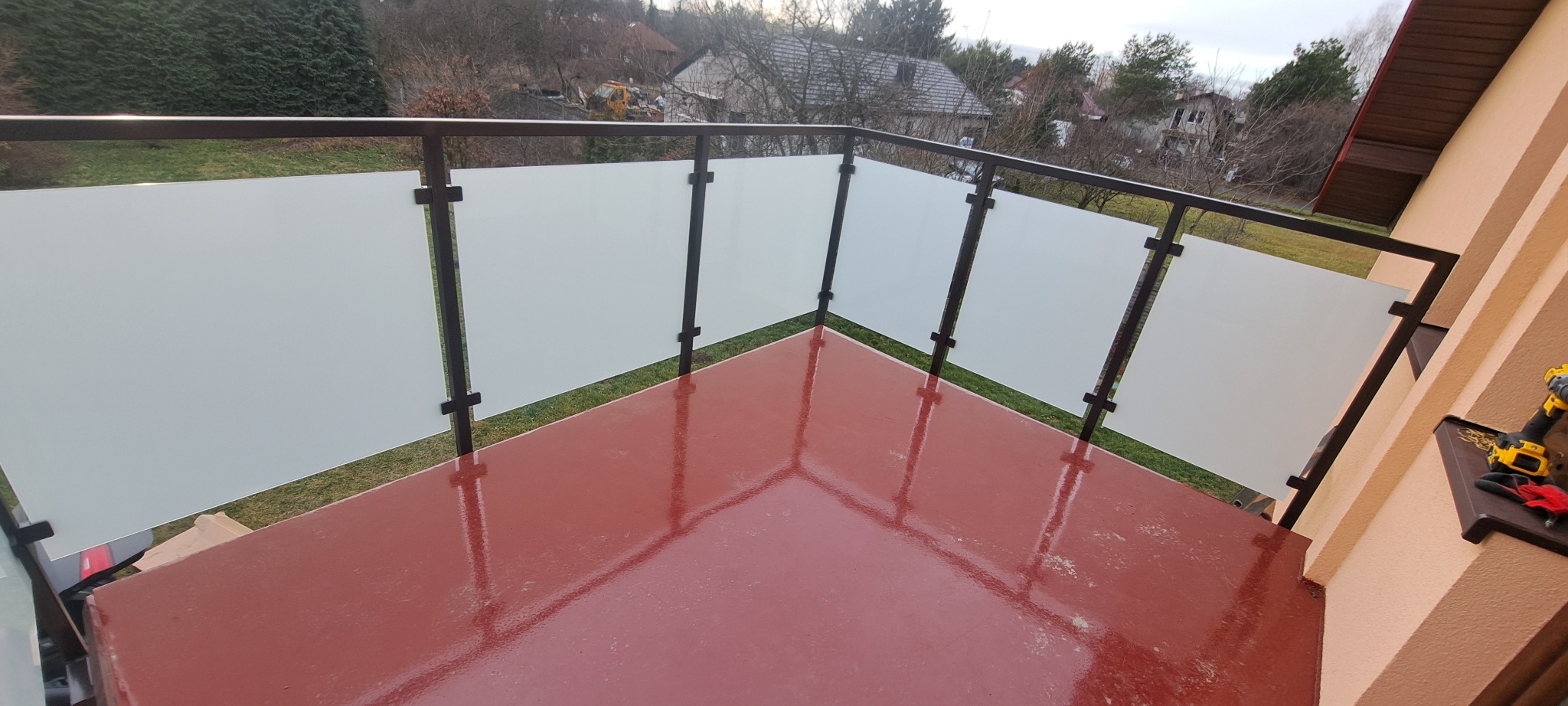 Balkon z nowoczesną balustradą ze szkła mlecznego i ciemnobrązowych profili, widok z góry na czerwoną posadzkę, narzędzia monterskie w rogu.