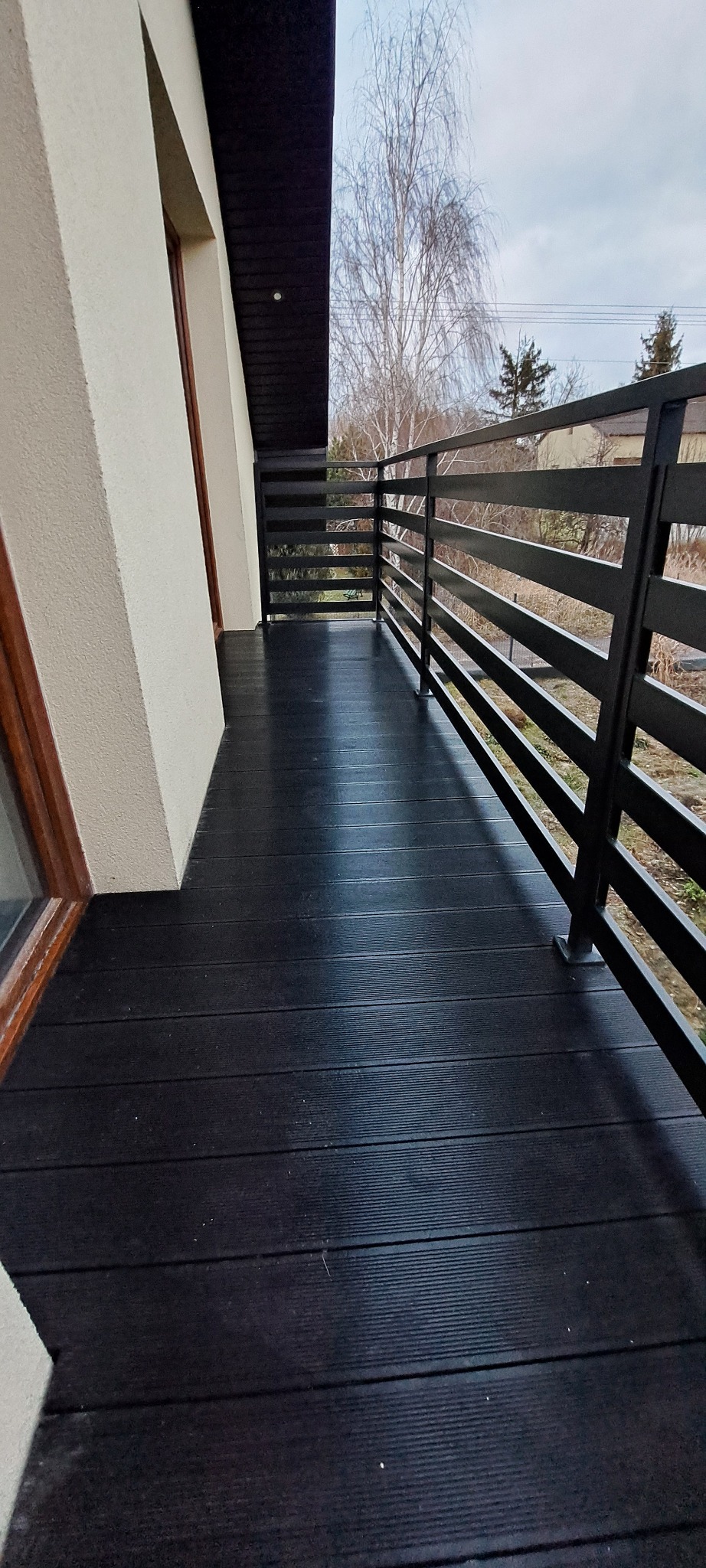 Czarny balkon z poziomymi, metalowymi barierkami i deskami podłogowymi, widok na drzewa bez liści w tle.