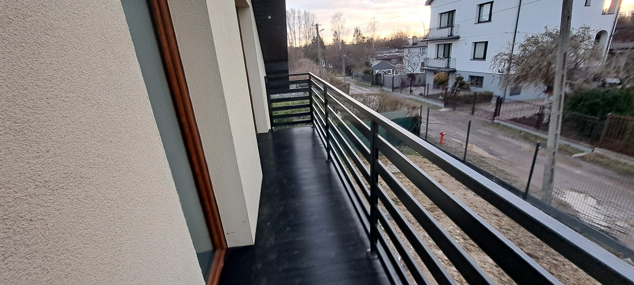 Balkon z prostą, czarną balustradą w Aleksandrowie Łódzkim, widok na ulicę i sąsiednie budynki o zmierzchu.