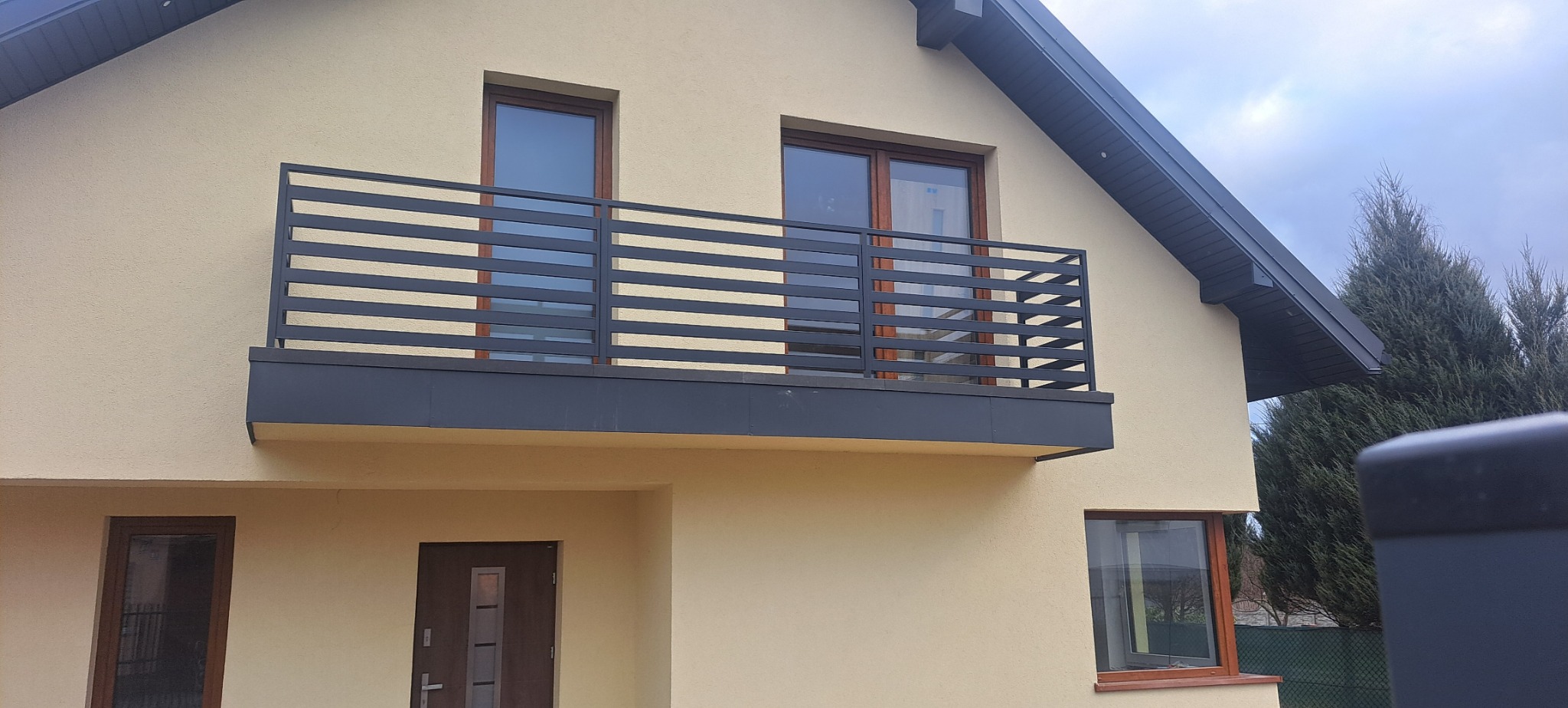 Nowoczesna balustrada balkonowa z poziomych, ciemnych profili na tle jasnej elewacji domu z drewnianą stolarką okienną i ciemnym dachem.