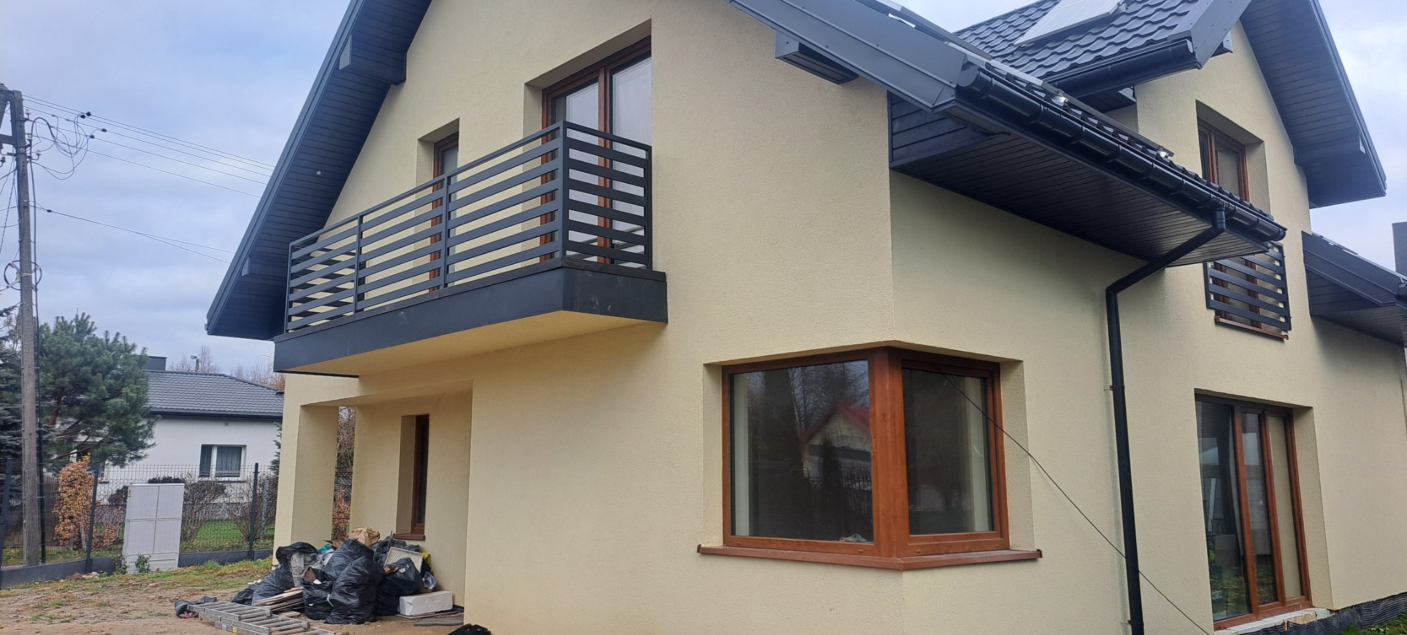 Nowoczesny dom z beżową elewacją i balkonem z czarną, poziomą konstrukcją, widoczne okna z drewnianymi ramami, rynny i orynnowanie w kolorze grafitowym, na dole stos worków ze śmieciami i drabina.