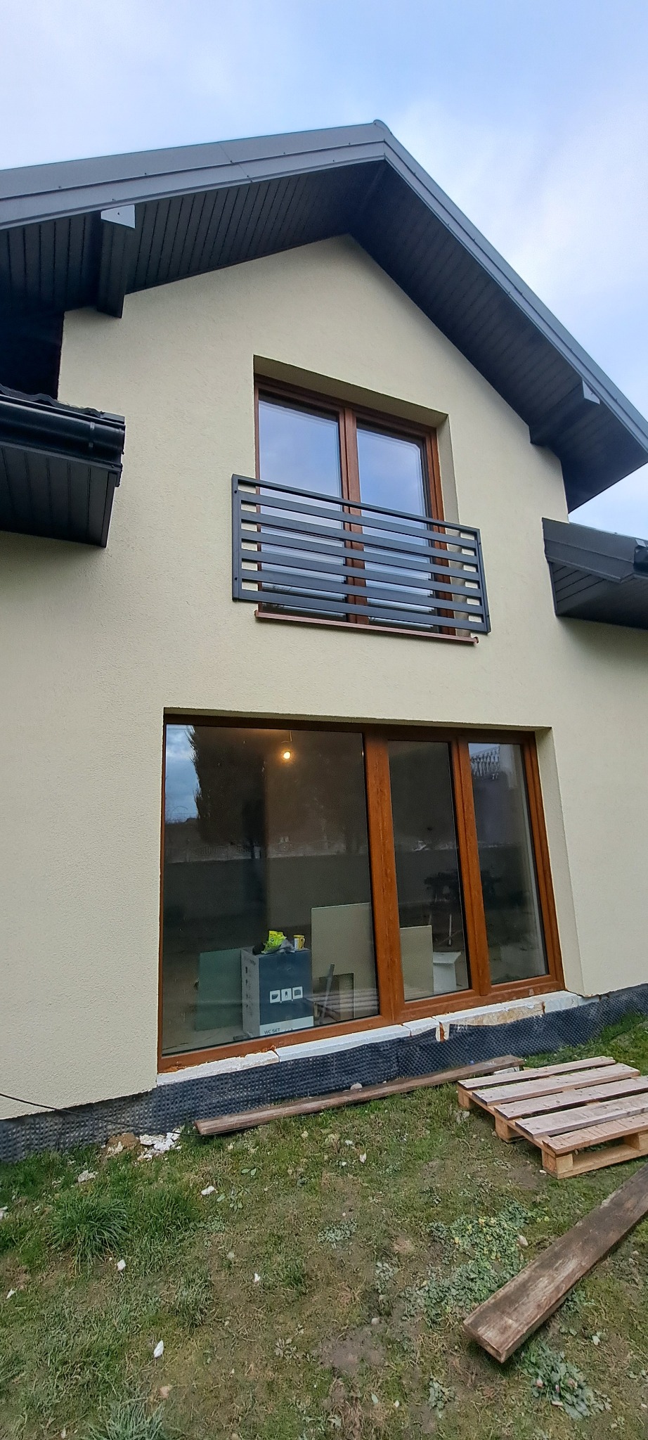 Elewacja domu z oknem balkonowym zabezpieczonym czarną, poziomą konstrukcją. Na parterze duże okno z drewnianą ramą, wewnątrz widoczne materiały budowlane. Przed domem trawa i palety.