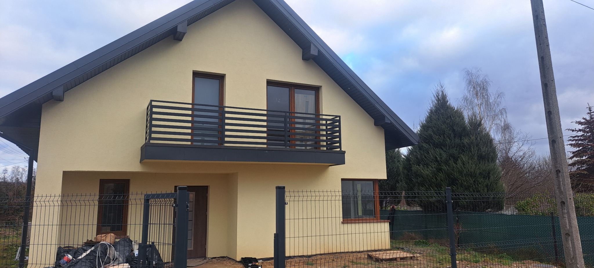 Nowoczesny dom jednorodzinny z beżową elewacją, balkonem z poziomą, czarną konstrukcją, otoczony siatką ogrodzeniową w trakcie montażu, z widocznymi workami z materiałami budowlanymi.
